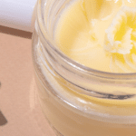 A glass jar filled with a creamy, yellow balm-like substance, resembling beef tallow, placed on a neutral surface with skincare tools nearby.