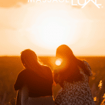 Two people sitting together on a blanket in an open field during sunset, enjoying a calm moment outdoors.