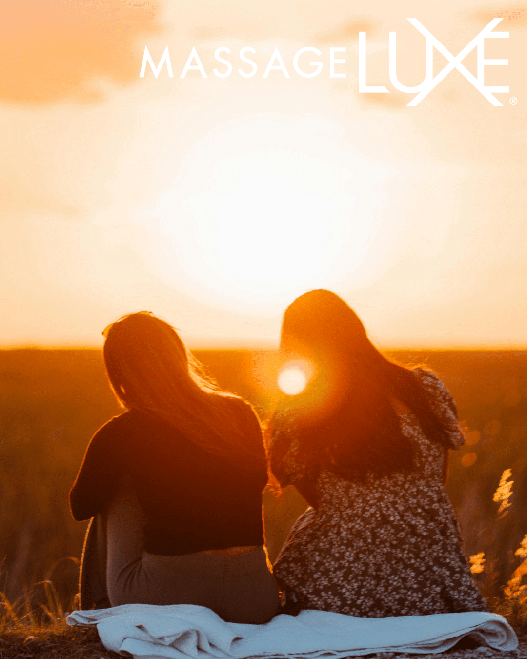 Two people sitting together on a blanket in an open field during sunset, enjoying a calm moment outdoors.