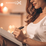 Woman writing in a journal beside soft candlelight in a calm, cozy setting. MassageLuXe logo displayed in the top corner.