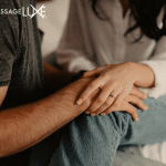 Close-up of a couple sitting together with one partner gently holding the other’s arm, creating a calm and supportive moment. MassageLuXe logo displayed in the top corner.