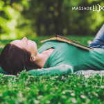 A woman lies peacefully on a blanket in a grassy park with a book resting on her chest, enjoying a quiet moment of relaxation outdoors.