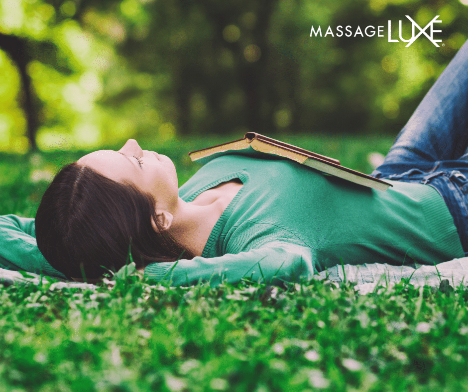 A woman lies peacefully on a blanket in a grassy park with a book resting on her chest, enjoying a quiet moment of relaxation outdoors.